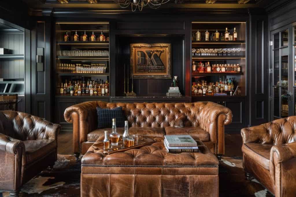 Sophisticated home whiskey lounge with deep-buttoned leather sofas, a central tufted ottoman, and dark wooden cabinetry housing an extensive whiskey collection.