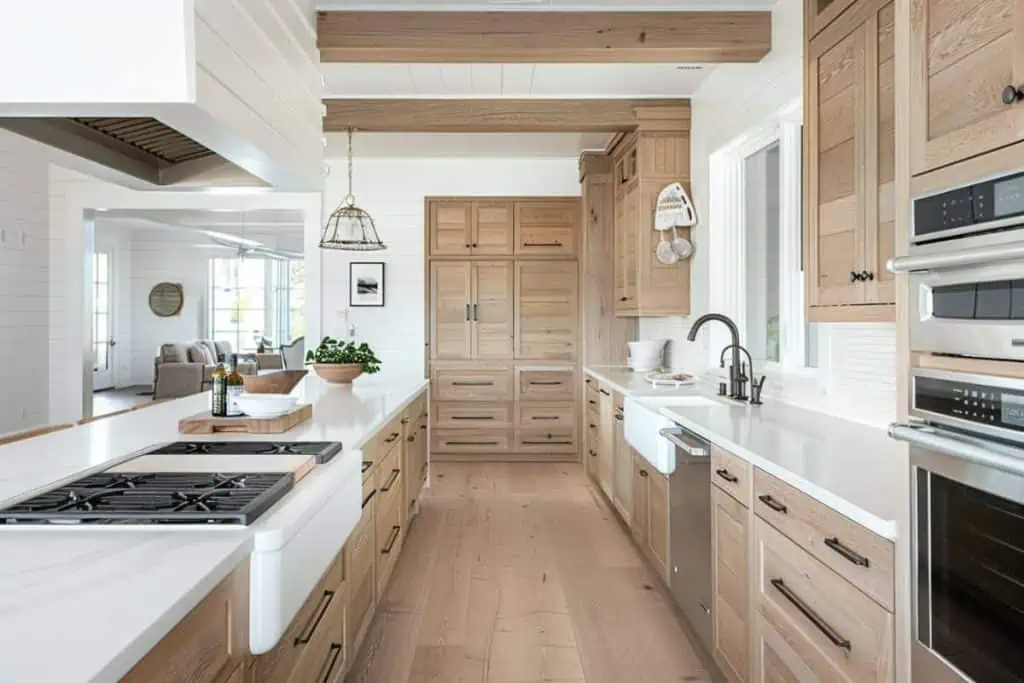 Open white kitchen with white oak cabinets, a central island, and pendant lighting, adjacent to a comfortable living area.