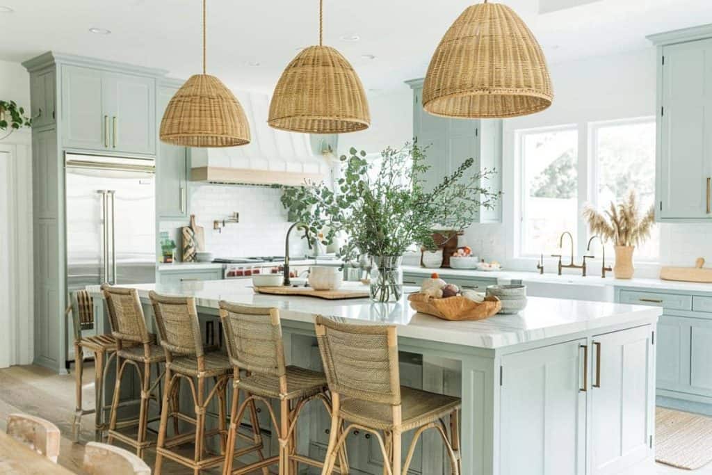 A sunlit coastal farmhouse kitchen with a serene palette, featuring three large wicker dome pendant lights over a spacious island with rattan bar stools, complemented by soft green cabinetry and fresh greenery in a large vase.