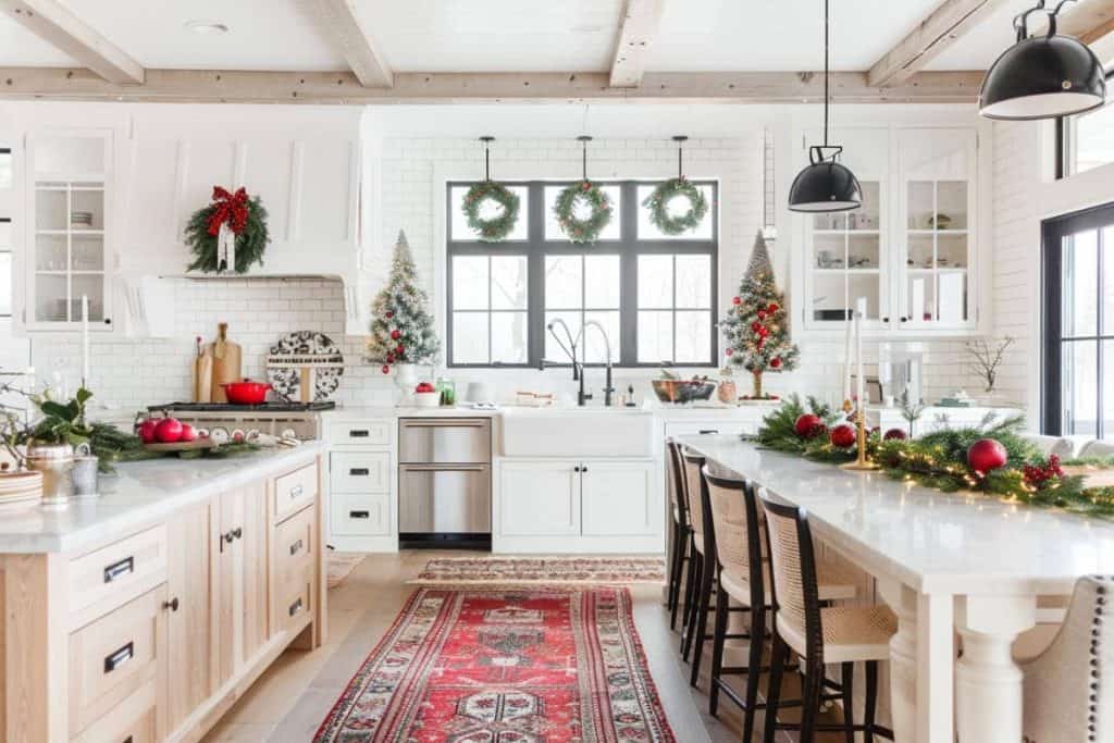 Charming farmhouse kitchen decorated for Christmas, showcasing a red festive rug, green wreaths on the window, and a large island with a garland centerpiece.