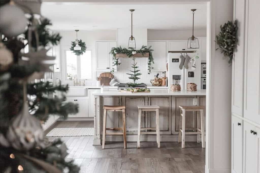 Rustic farmhouse kitchen with white island, wooden bar chairs, festive decorations, and a snow-flecked Christmas tree.