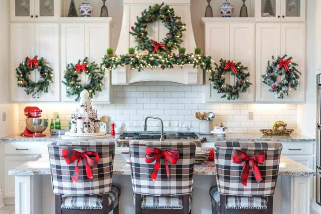 A luxurious farmhouse kitchen with white cabinets and a large island, decorated with garlands and wreaths, with plaid ribbon-tied chairs ready for a festive gathering.