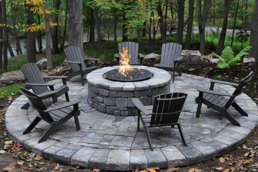 Elegant backyard fire pit area with gray Adirondack chairs on a circular stone patio, with a forest backdrop and fallen leaves scattered around