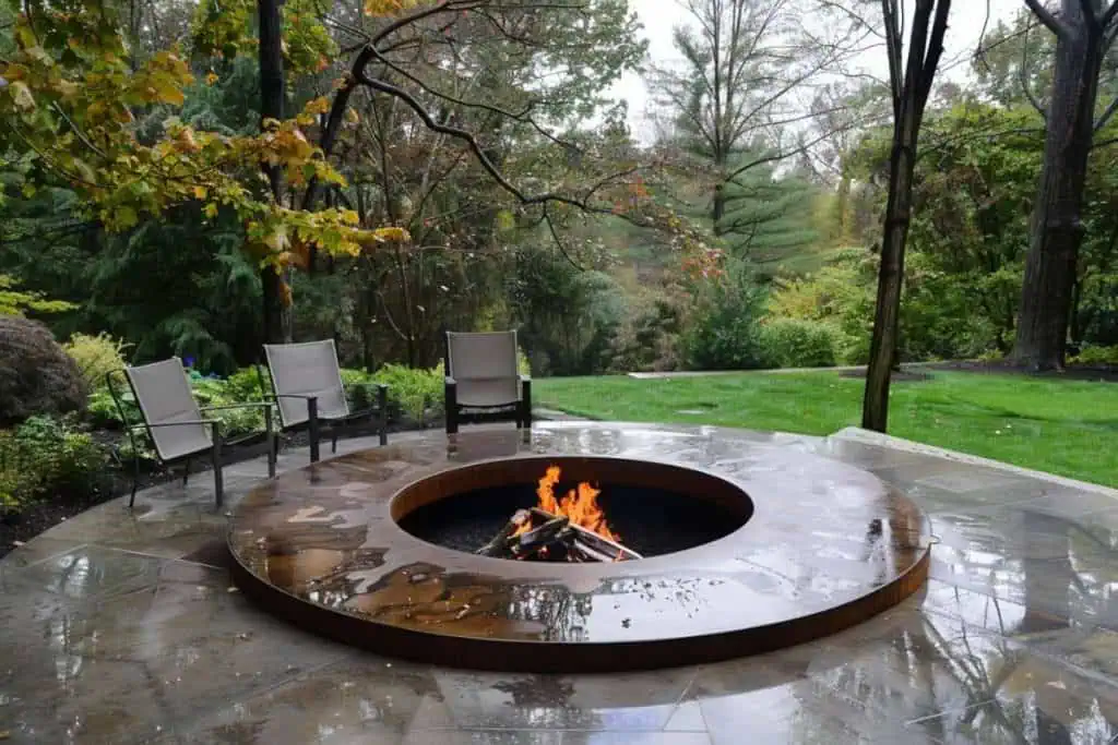 Modern backyard fire pit on a rainy day, with sleek chairs around it, glistening wet patio, and a lush, wooded backdrop