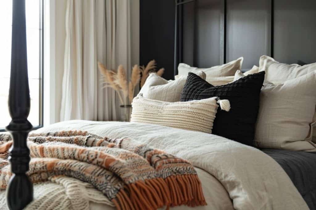 Detail of a black and neutral bedroom with textured pillows, a knit throw blanket in earthy tones, and a view of a window.