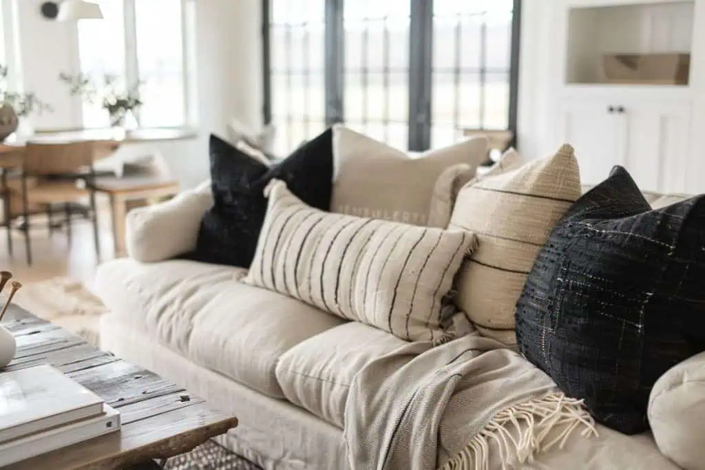 Detailed view of a black and neutral living room setting showing a cream-colored sofa with textured black, beige, and gray pillows, and a rustic wooden coffee table in the foreground.