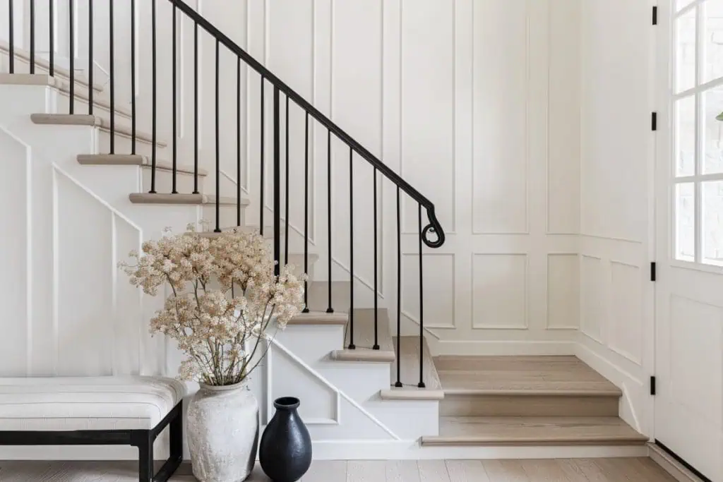 Elegant, minimalist entryway featuring a staircase with wooden steps and a black metal balustrade. A large vase with dried flowers sits on a bench beside a smaller black vase, complementing the white paneled walls and the light wooden flooring