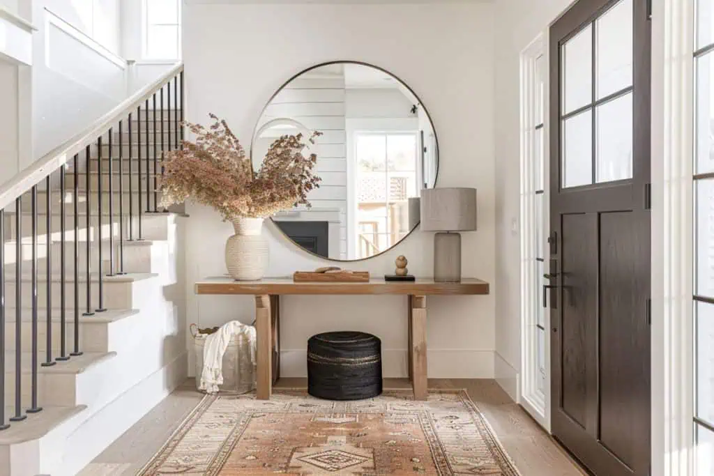 An inviting entryway featuring a wooden bench with a neutral cushion next to a large circular mirror. A vase of dried pink flowers adds a touch of nature, complementing the simple, modern decor