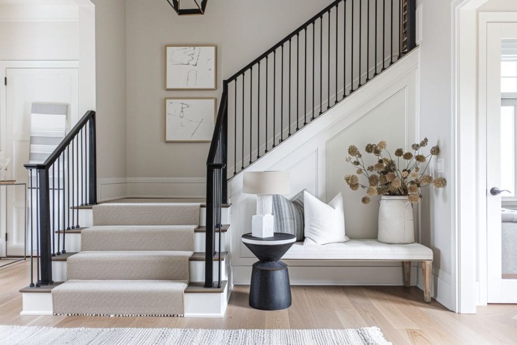 Modern entryway with a black staircase and matching balusters. A stylish bench with neutral upholstery sits beneath a collection of abstract artwork, paired with a decorative vase of dried flowers