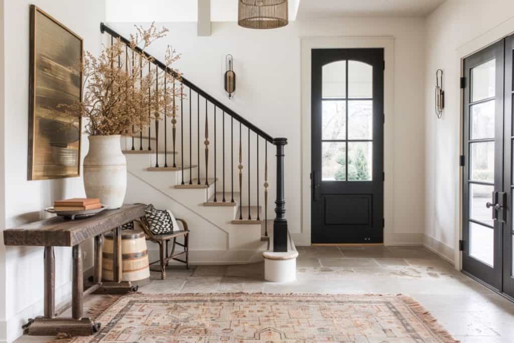 Rustic entryway with a dark wood bench and a matching staircase with ornate metal balusters. The space is accented with a large potted plant and a traditional rug, creating a warm, inviting atmosphere.