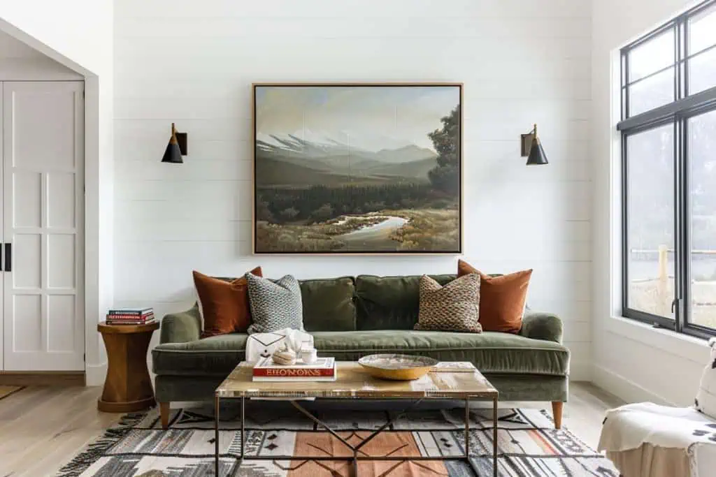 Rustic living room featuring a green velvet sofa, wooden ceiling beams, and large windows providing ample natural light. The room is decorated with a mix of traditional and contemporary elements, including a large painting of a mountain landscape.
