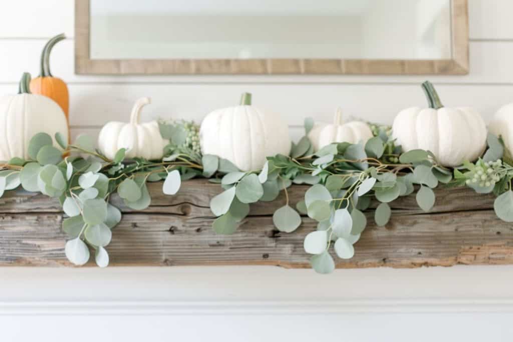 A wooden mantle decorated with white pumpkins, green foliage, and a variety of white candles, presenting a calm and peaceful fall decor.