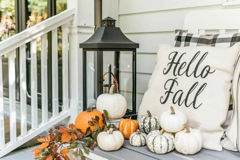 A cozy porch decorated for fall with a pillow reading "Hello Fall," surrounded by various white and orange pumpkins. A black lantern and autumn leaves complete the setting.