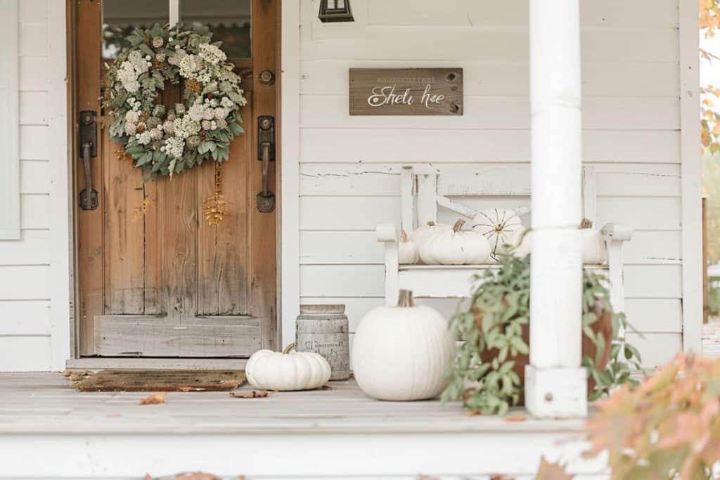 A wooden front door with a large dried floral wreath, surrounded by white pumpkins and a wooden bench