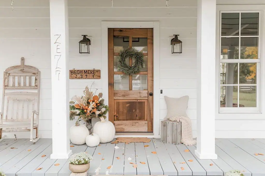 A porch with a wooden front door and a wreath, decorated with white pumpkins and a floral arrangement in a vase. A rustic rocking chair and wooden crate enhance the cozy fall decor.