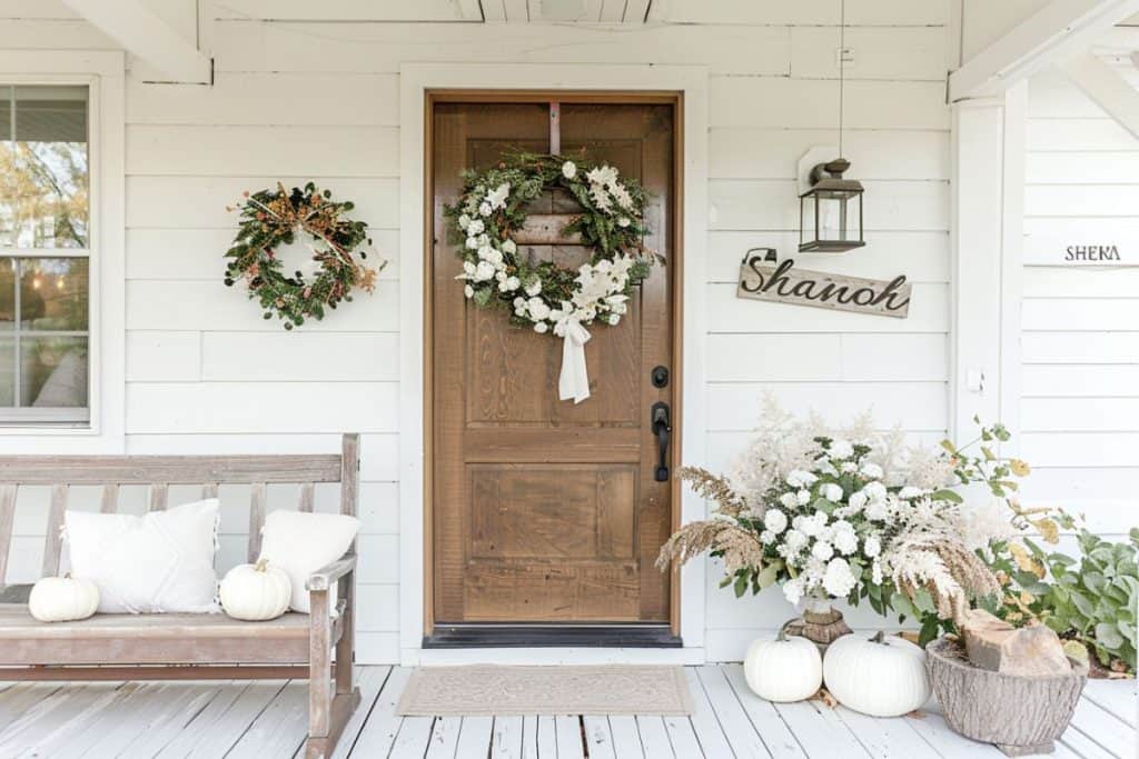 A front door with a lush green and white floral wreath. The porch features a wooden bench with white pumpkins and pillows, alongside a large floral arrangement