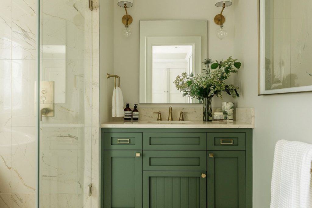 A sophisticated bathroom featuring a green vanity with brass hardware, a beige countertop, and a large mirror. The space includes a glass-enclosed shower with marble tiles and a floral arrangement on the counter.
