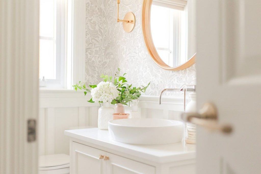 A bright bathroom with a white vanity, round mirror, and a vase of white flowers. The walls have intricate, floral-patterned wallpaper, and natural light streams in through the window.