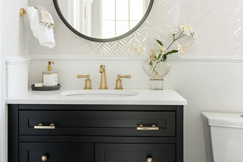 A modern bathroom featuring a black vanity with brass hardware, a round mirror, and a clear vase of white flowers. The backsplash has a chevron pattern, adding texture to the space.