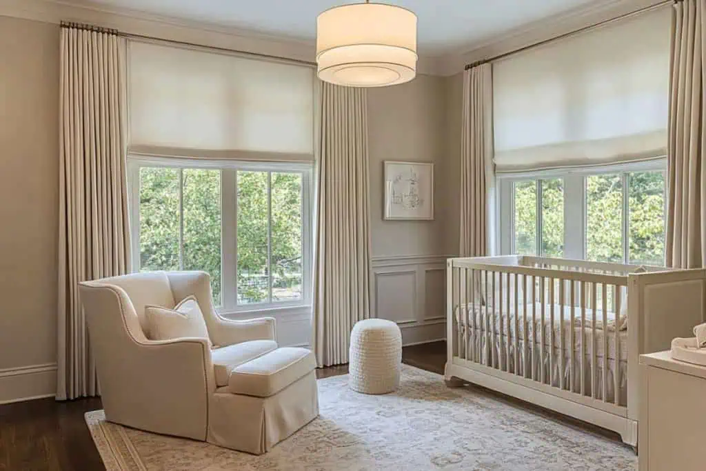A sophisticated nursery featuring cream curtains, a white crib, and an upholstered armchair. The room is filled with soft natural light, showcasing minimal wall decor and elegant, understated furnishings.