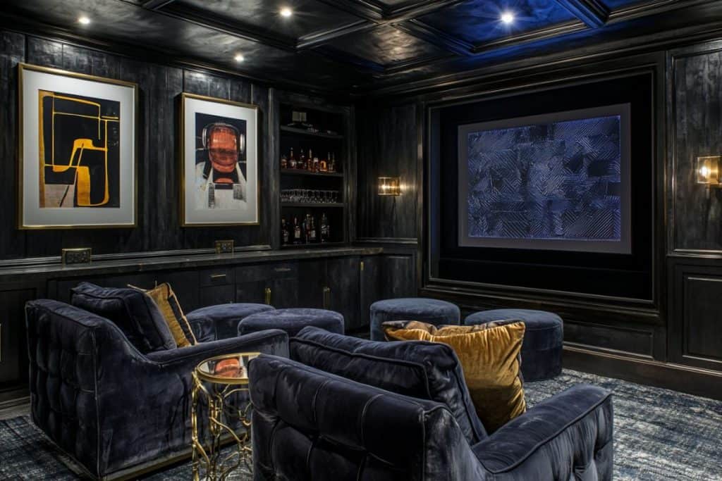 A cinematic man cave with plush navy blue velvet seating, a large screen, and dim ambient lighting. A mini-bar is stocked with premium spirits, and modern artwork decorates the dark walls.