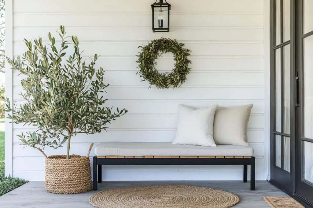 A simple yet elegant spring porch decor design with a woven bench, neutral cushions, and a potted olive tree in a woven basket. A circular woven rug adds texture, while a black-framed glass door enhances the modern, natural vibe.