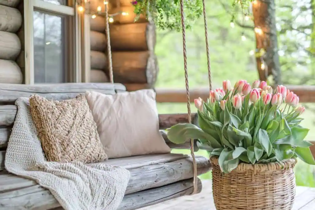 A cozy spring porch decor scene on a log cabin-style porch with a rustic hanging swing covered in neutral pillows and blankets. A woven basket overflowing with pink tulips sits beside the swing, adding a fresh floral touch. String lights above enhance the warm and inviting feel.